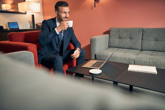 Tranquilo joven apuesto en un traje sentado con un teléfono inteligente y una computadora portátil en una cómoda zona de salón mientras toma café