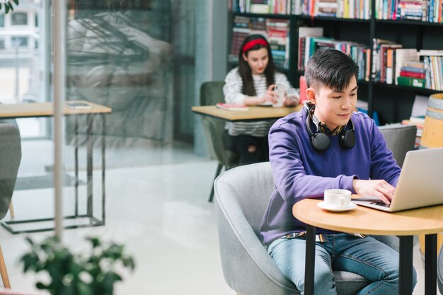 Tranquilo joven asiático usando una computadora portátil moderna mientras está sentado con una taza de café en la mesa pequeña