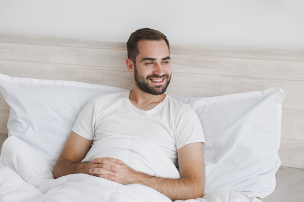 Foto tranquilo joven guapo barbudo acostado en la cama con sábana blanca manta de almohada en dormitorio en casa