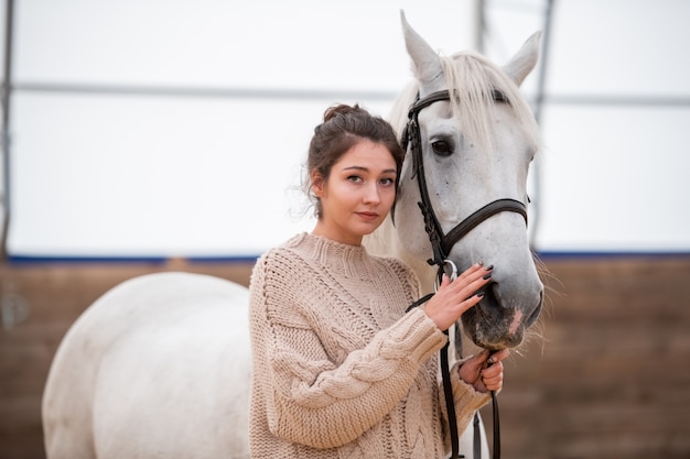 Tranquilo joven mujer casual en suéter de punto mirándote mientras está de pie junto al caballo de pura raza blanca en el establo