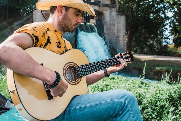 Tranquilo joven con un sombrero de vaquero tocando la guitarra en un área natural