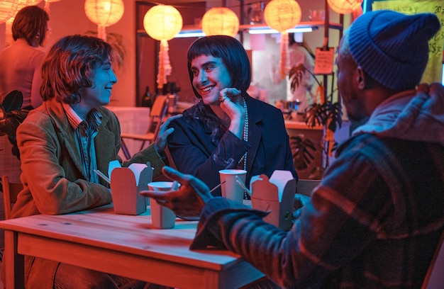 Transgénero sentado a la mesa con sus amigos y hablando con ellos durante la reunión en el bar.