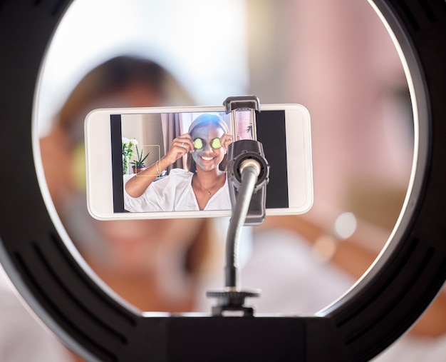 Transmisión en vivo de teléfono y mujer en anillo de luz para tutorial de cuidado de la piel o publicación en redes sociales en casa Sonrisa de bloguera feliz con máscara facial de belleza para vlog de Internet o podcast móvil