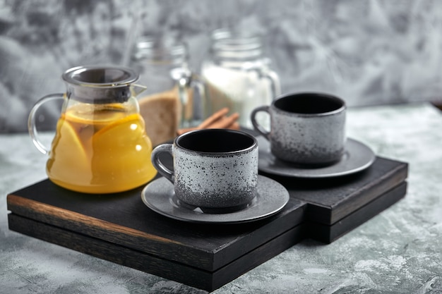 Transparente Glasteekanne mit Zitrusfruchttee und Schalen, Teesatz auf einem Holztisch. Close up, grau, weiches Licht.