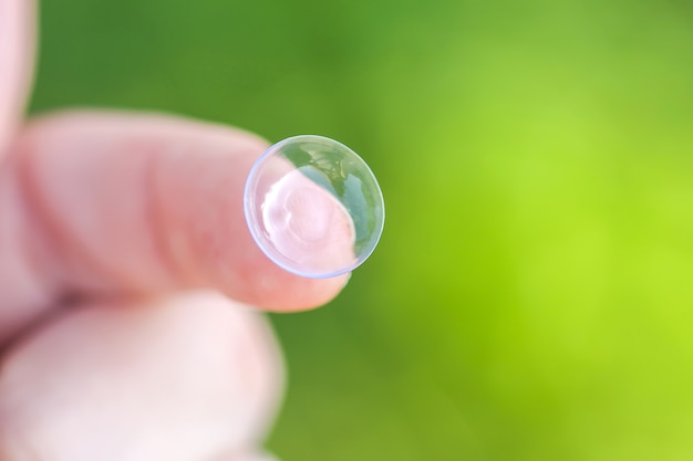 Transparente Kontaktlinse an der Fingerspitze auf unscharfem grünem Sommernaturhintergrund.