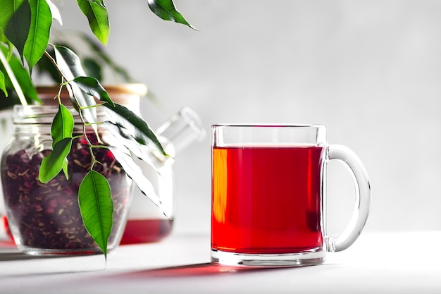 Transparenter Becher mit rotem Tee auf grauem Hintergrund, neben einem Glas mit Hibiskus und einer Teekanne