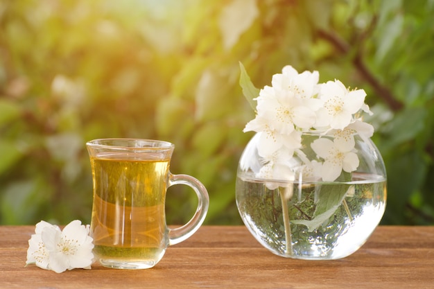 Transparenter Becher Tee und ein Vase mit Jasmin auf einem Holztisch, Grüns