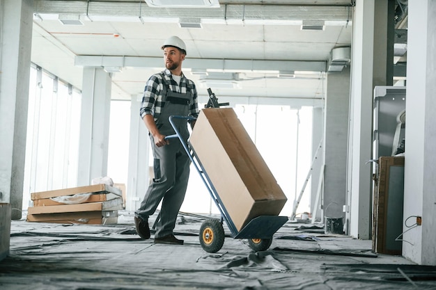 Transportando una larga caja de papel Trabajador de la construcción en uniforme en una habitación vacía sin terminar
