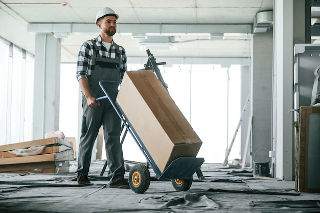 Transportando una larga caja de papel Trabajador de la construcción en uniforme en una habitación vacía sin terminar