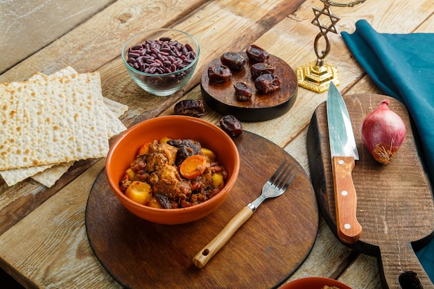 Transporte de prato judaico com carne em um prato sobre uma mesa de madeira ao lado de pão ázimo. Garfo e ingredientes.