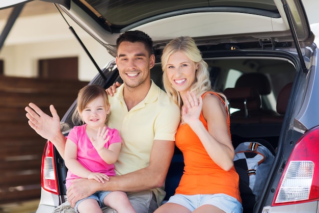 transporte, lazer, viagem e conceito de pessoas - família feliz com menina sentada no porta-malas do carro hatchback e acenando as mãos em casa espaço de estacionamento