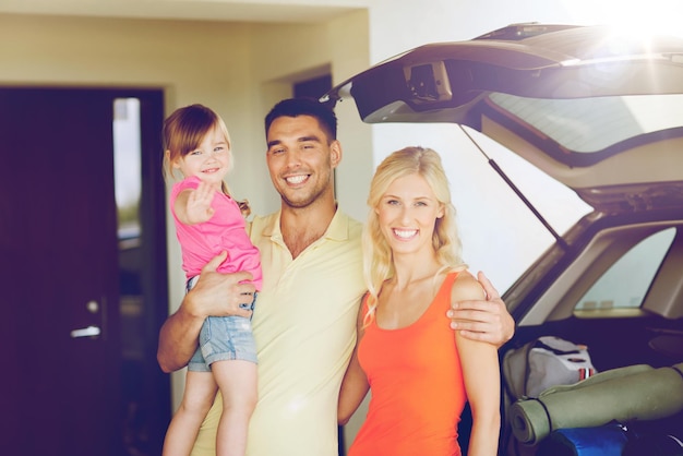 transporte, ocio, viaje por carretera, viajes y concepto de personas - familia feliz con una niña pequeña empacando cosas en un auto con puerta trasera en el estacionamiento de su casa