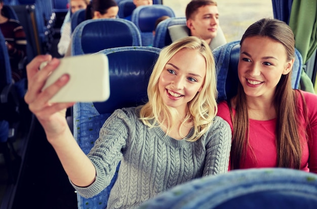 transporte, turismo, viaje por carretera y concepto de personas - mujeres jóvenes felices o amigas en autobús de viaje tomando selfie por teléfono inteligente