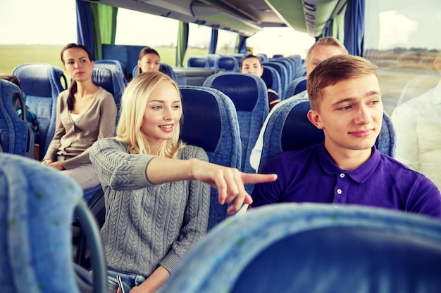 transporte, turismo, viaje por carretera y concepto de personas - pareja joven con un grupo de turistas conduciendo en autobús de viaje y señalando con el dedo algo fuera