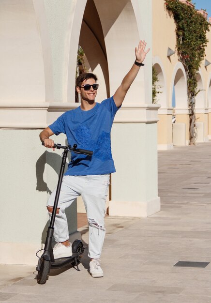 Transporte urbano. Chico joven feliz en ropa casual con scooter eléctrico en la calle de la ciudad, saludando a alguien, saludando a un amigo al aire libre, copie el espacio. Ocio activo, concepto de deportes de verano