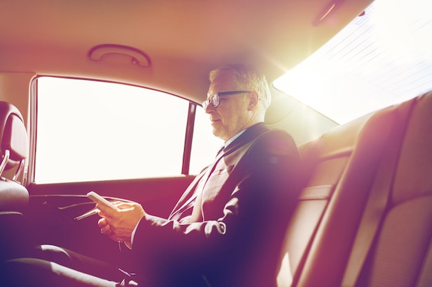 transporte, viaje de negocios, tecnología y concepto de personas - hombre de negocios senior enviando mensajes de texto en un teléfono inteligente y conduciendo en el asiento trasero del camión
