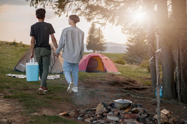 Traseira de casal caminhando em acampamento de mãos dadas e carregando geladeira portátil