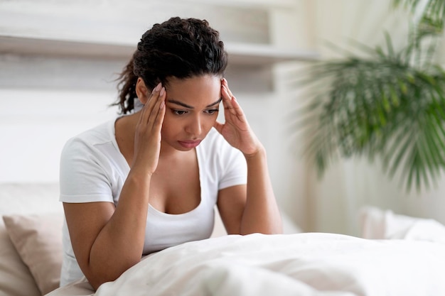 Los trastornos del sueño molestan a la mujer negra sentada en la cama y tocando la cabeza de cerca