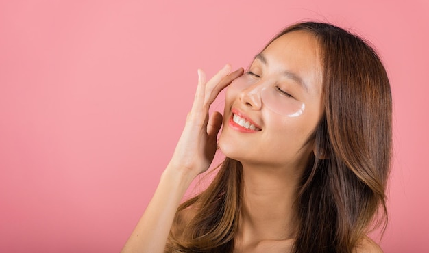 Tratamento da pele dos olhos. Uma jovem asiática bonita e sorridente máscara cosmética gel sob os olhos, Retrato feminino com maquiagem natural hidrogel tapa-olho no rosto, isolado em fundo rosa, cuidados com a pele