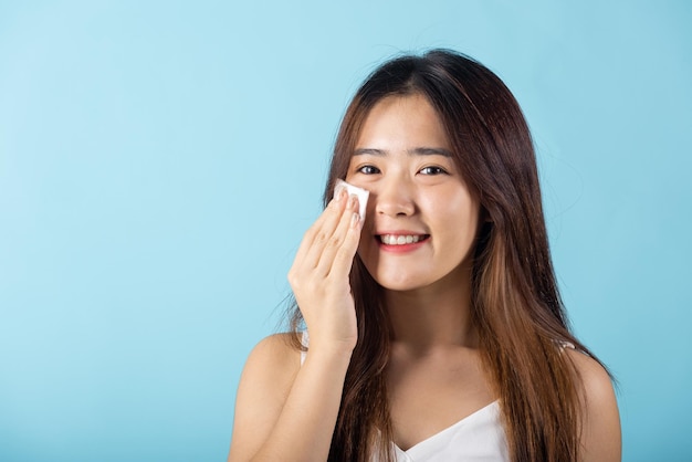 Tratamento de Beleza Facial Higiene da pele. Mulher jovem e bonita asiática removendo maquiagem cosmética usando disco de limpeza facial de algodão, tiro de estúdio isolado em fundo azul, olhos de limpeza femininos felizes