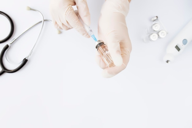 Tratamento medicamentoso, conceito de vacinação. Médico mãos segurando um frasco com vacina e seringa de injeção, termômetro eletrônico, estetoscópio e frasco de vacina em fundo branco.
