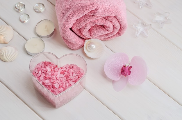 Tratamentos de spa como presente de Dia dos Namorados. Toalha rosa com flor, conchas e sal marinho rosa em forma de coração sobre uma superfície de madeira branca. Salão de beleza, massagem.