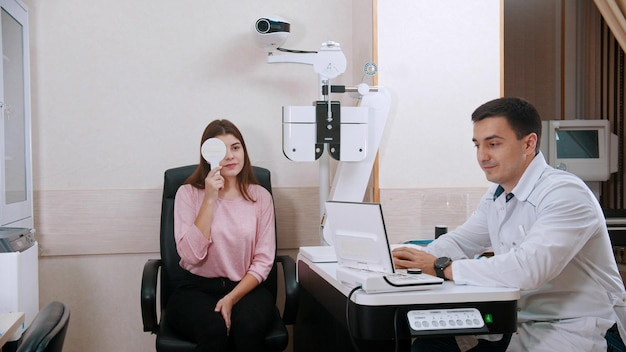 Tratamiento oftalmológico en el gabinete mujer joven comprobando su agudeza visual cerrando el ojo con un protector ocular