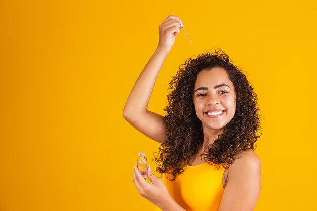 Tratamiento reparador de puntas abiertas. Sonriente mujer africana rociando aceite esencial en su cabello castaño rizado