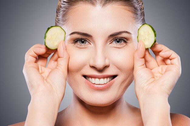 Foto tratamiento de spa natural. hermosa mujer madura sosteniendo trozos de pepino cerca de sus ojos y sonriendo mientras está de pie aislado sobre fondo blanco.