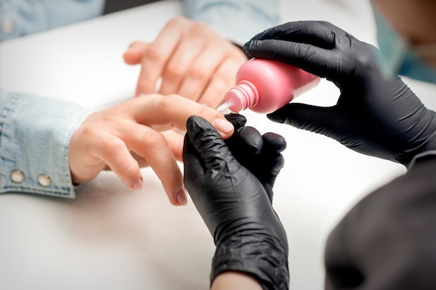 Tratamiento de uñas con aceite para cutículas mientras manicura de mujer joven en salón de uñas