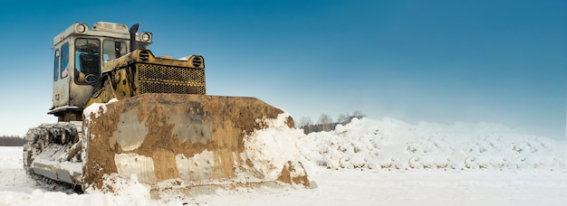 Trator de esteira amarela com um balde funciona no inverno, limpando a estrada da neve.