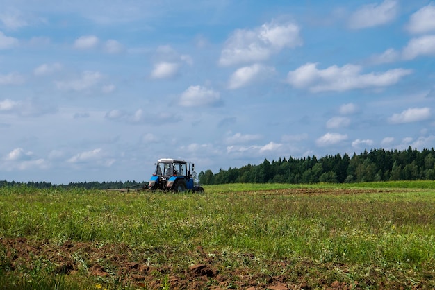 Trator trabalhando no campo arando a terra preparando-se para a semeadura vista do campo e trator à distância contra um céu azul com nuvens