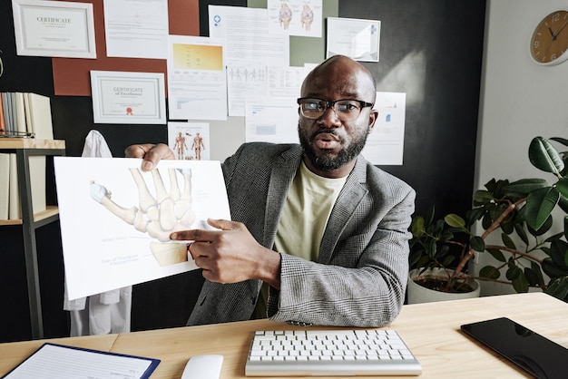 Traumatologista masculino africano apontando para a foto na mão durante a conferência online no computador em