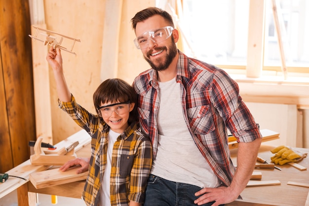 Traumschöpfer. Fröhlicher kleiner Junge, der ein Holzflugzeug hält, während er mit seinem Vater in der Werkstatt steht