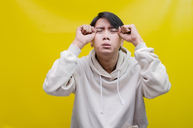 Traurig Ein junger asiatischer Mann, der ein graues T-Shirt mit weinendem Gesichtsausdruck trägt, ist isoliert auf Gelb