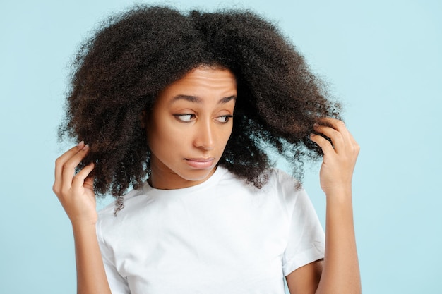 Traurige afroamerikanische Frau mit lockigem Haar berührt ihr Haar, Haarausfall, isoliert auf blauem Hintergrund