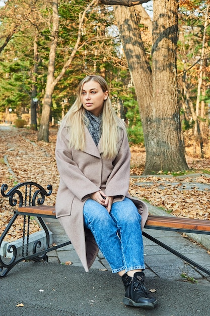 Traurige blonde Frau im grauen Mantel sitzt auf einer Parkbank