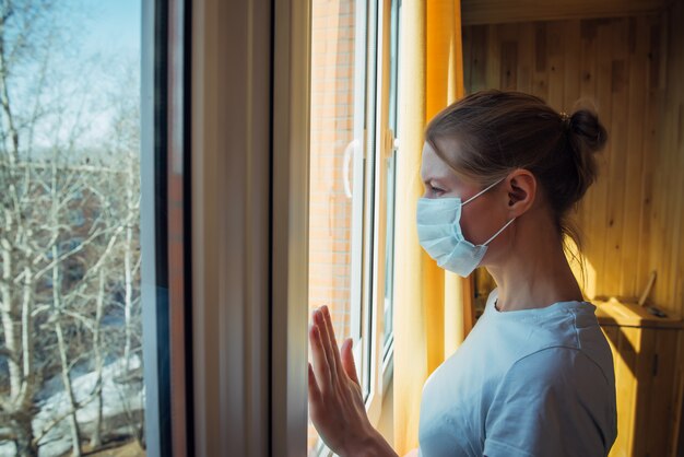 Traurige einsame Frau in der schützenden medizinischen Maske auf ihrem Gesicht, das Fenster betrachtet. Chinesisches Pandemie-Coronavirus, Virus Covid-19. Quarantäne, Infektionsprävention, Isolierung zu Hause, Schutz gegen SARS-CoV-2