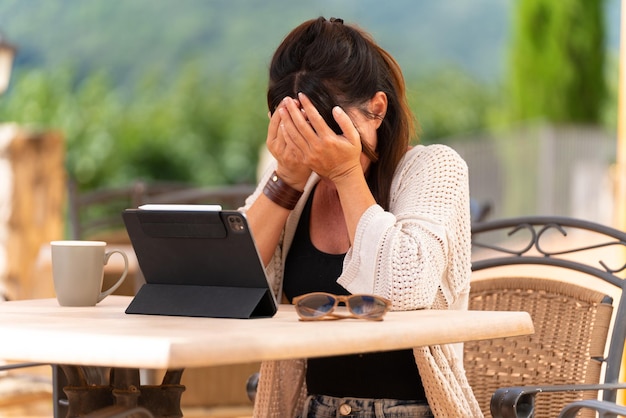 Traurige Frau weint an einem Tisch auf einer Terrasse im Freien mit einem Tablet mit den Händen im Gesicht