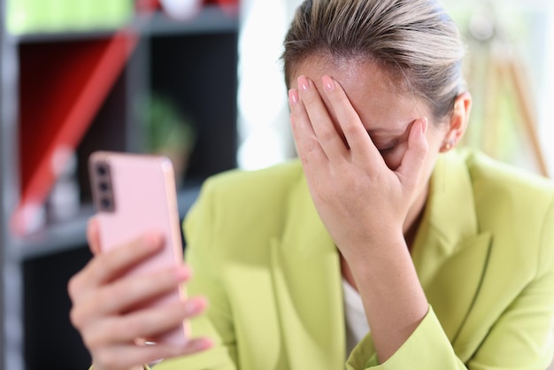 Traurige Geschäftsfrau hält Smartphone in der Hand und bedeckt ihr Gesicht mit der Hand