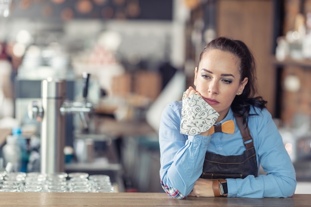 Traurige Geschäftsinhaberin hält Gesichtsmaske in der Hand und lehnt sich in ihrem leeren Café an die Bar.