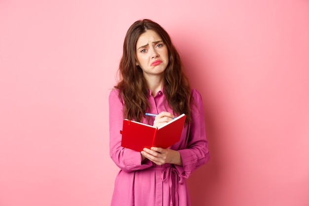 Traurige junge Frau, die ihre Gefühle in Tagebuch schreibt, verärgert und düster aussieht, sich gebrochen fühlt, Erinnerungen im Planer teilt, gegen rosa Wand steht.