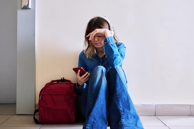 Traurige junge Studentin mit Rucksack-Smartphone auf dem Boden sitzend