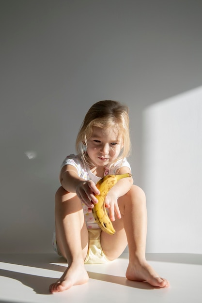 Traurige Kindermädchenblondine, die eine Banane hält, die auf dem Boden vor dem Hintergrund einer weißen Wand sitzt