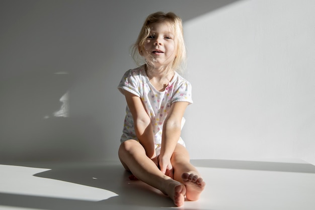 Traurige Kindermädchenblondine sitzt auf einem weißen Boden vor dem Hintergrund einer weißen Wand