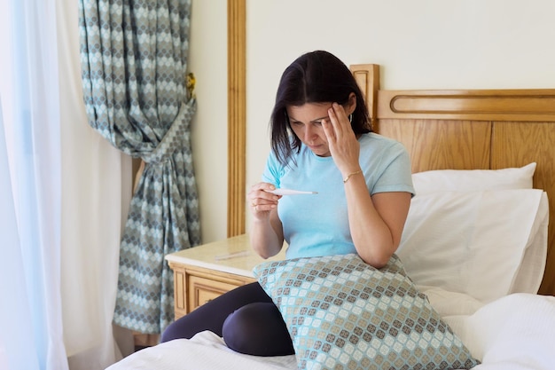 Traurige kranke reife Frau, die ein Thermometer in ihren Händen hält und zu Hause im Bett sitzt