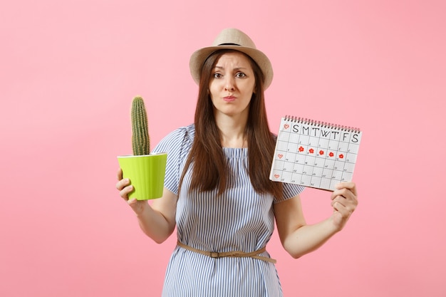 Traurige Krankheitsfrau im blauen Kleid mit grünem Kaktus, Periodenkalender zur Überprüfung der Menstruationstage einzeln auf rosafarbenem Hintergrund. Medizin, Gesundheitswesen, Gynäkologie, Bauchschmerzkonzept. Platz kopieren.