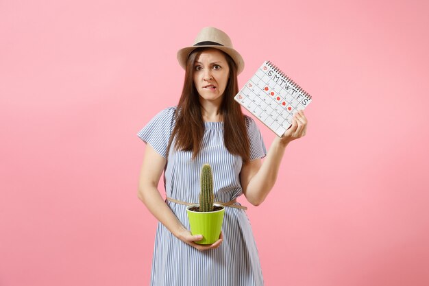 Traurige Krankheitsfrau im blauen Kleid mit grünem Kaktus, Periodenkalender zur Überprüfung der Menstruationstage einzeln auf rosafarbenem Hintergrund. Medizin, Gesundheitswesen, Gynäkologie, Bauchschmerzkonzept. Platz kopieren.