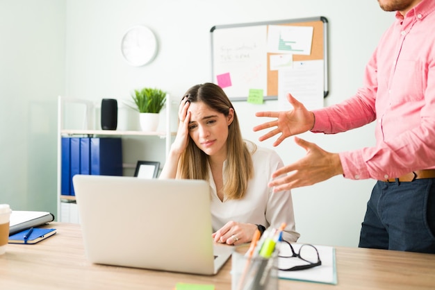 Traurige schöne Frau in ihren 20ern, die wegen eines Arbeitsfehlers das Geschrei und Geschrei ihres männlichen Chefs hört, während sie vor einem Laptop am Schreibtisch sitzt