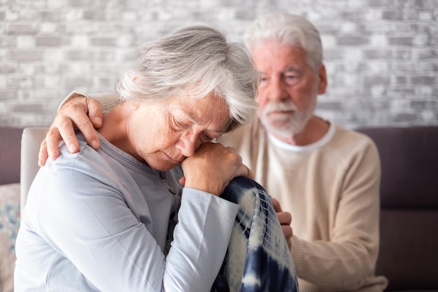 Traurige und depressive ältere Frau, die zu Hause sitzt, während der Ehemann versucht, ihren unglücklichen Senior zu trösten